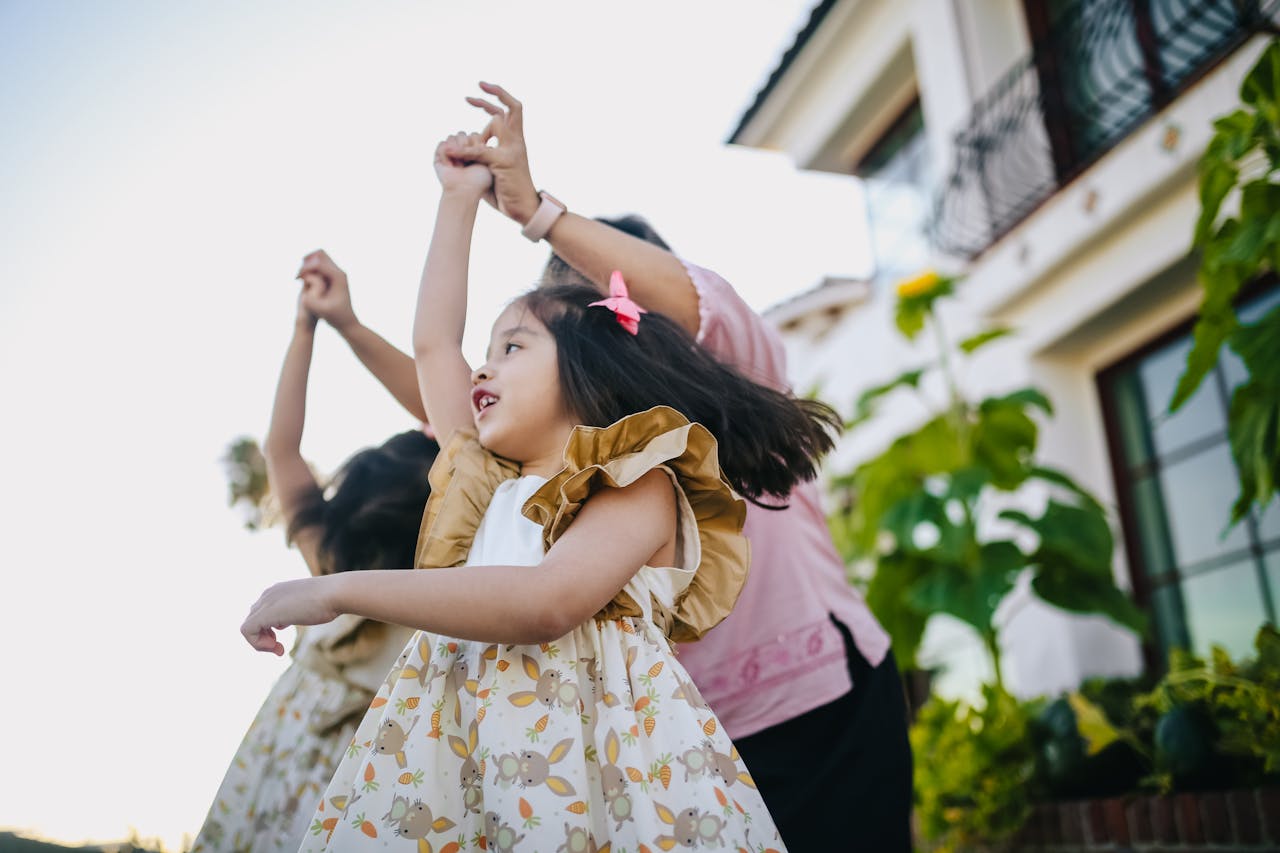 about-img A joyful moment of a mother and her daughters dancing and playing outside on a sunny day.