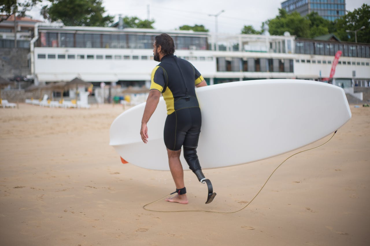 portfolio-img-04 A man with a prosthetic leg carries a surfboard on a beach in Portugal, symbolizing strength and adventure.